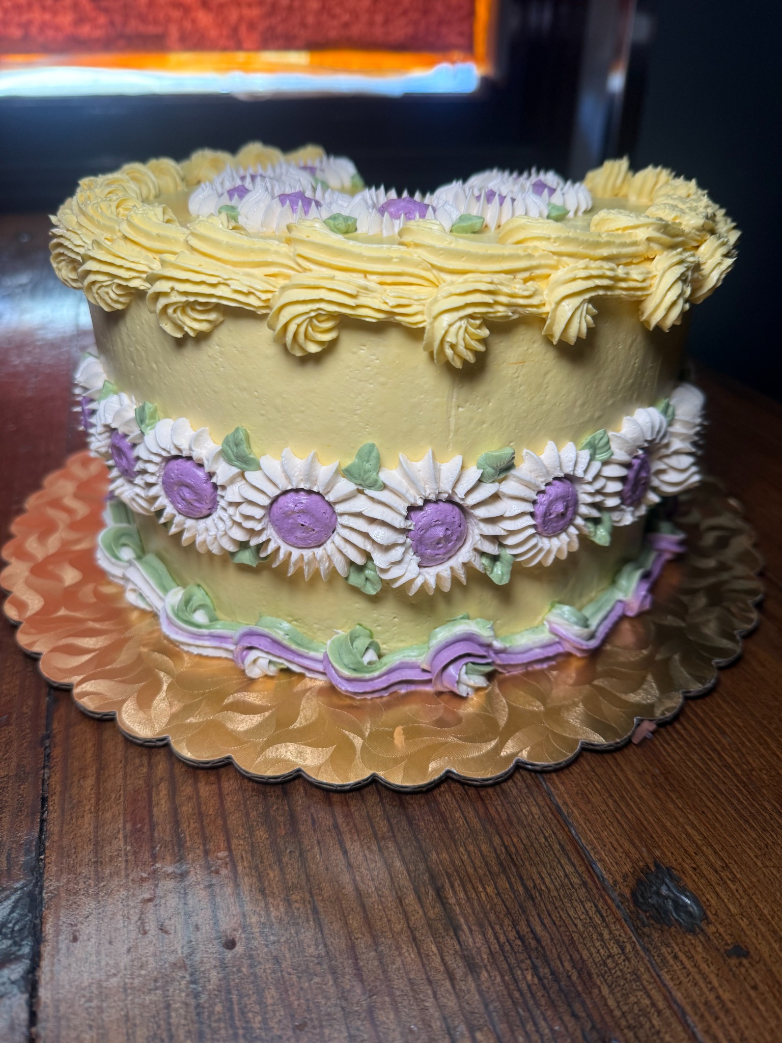 Yellow buttercream cake with white daisies, lavender centers, and green leaves